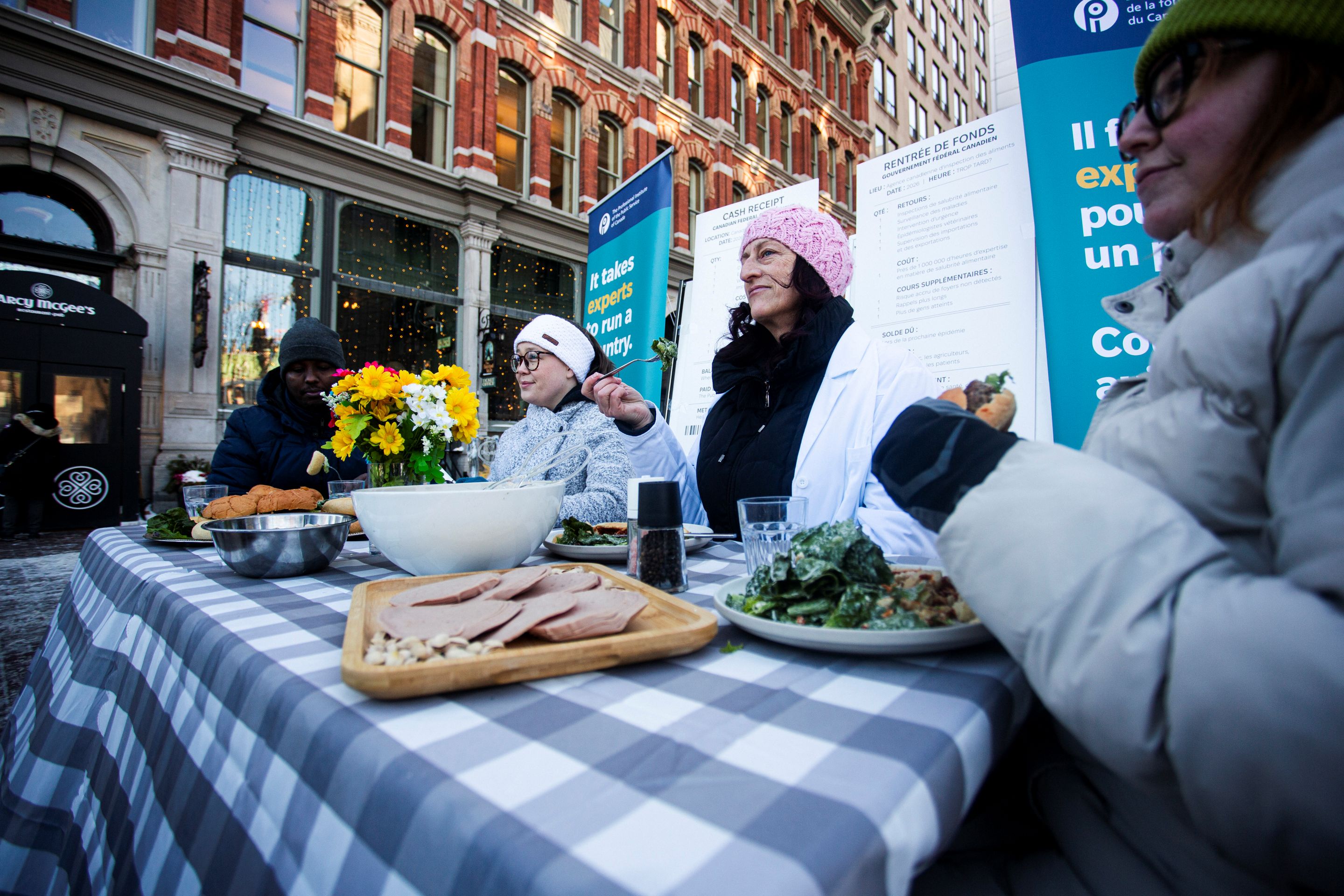 Avec en arrière-plan des personnes partageant un repas et une table garnie de produits alimentaires qui ont subi un rappel, les expert·es de la salubrité alimentaire avertissent aujourd’hui que les réductions du gouvernement fédéral dans l’Agence canadienne d’inspection des aliments (ACIA) affaiblissent la capacité du Canada à prévenir les maladies d’origine alimentaire.